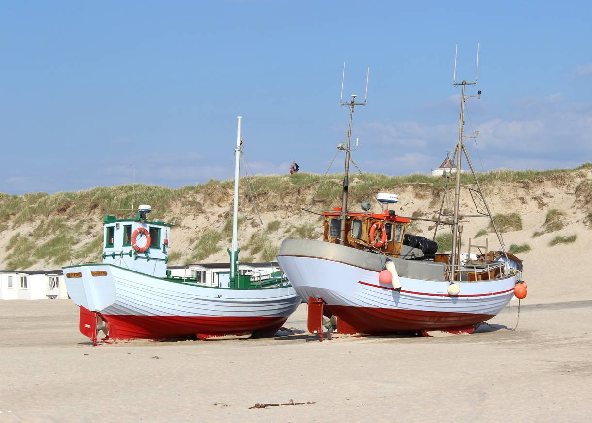 Hellblau-rotre Fischerboote auf dem Strand, dahinter Dünen und weiße Badehüttten