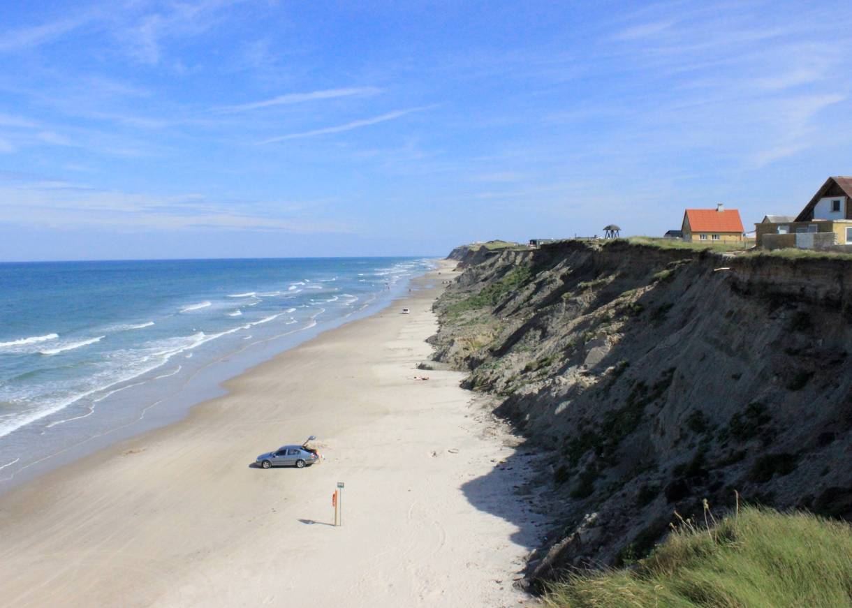 Steilküste im Meer, unten Sandstrand, oben stehen vereinzelte Häuser