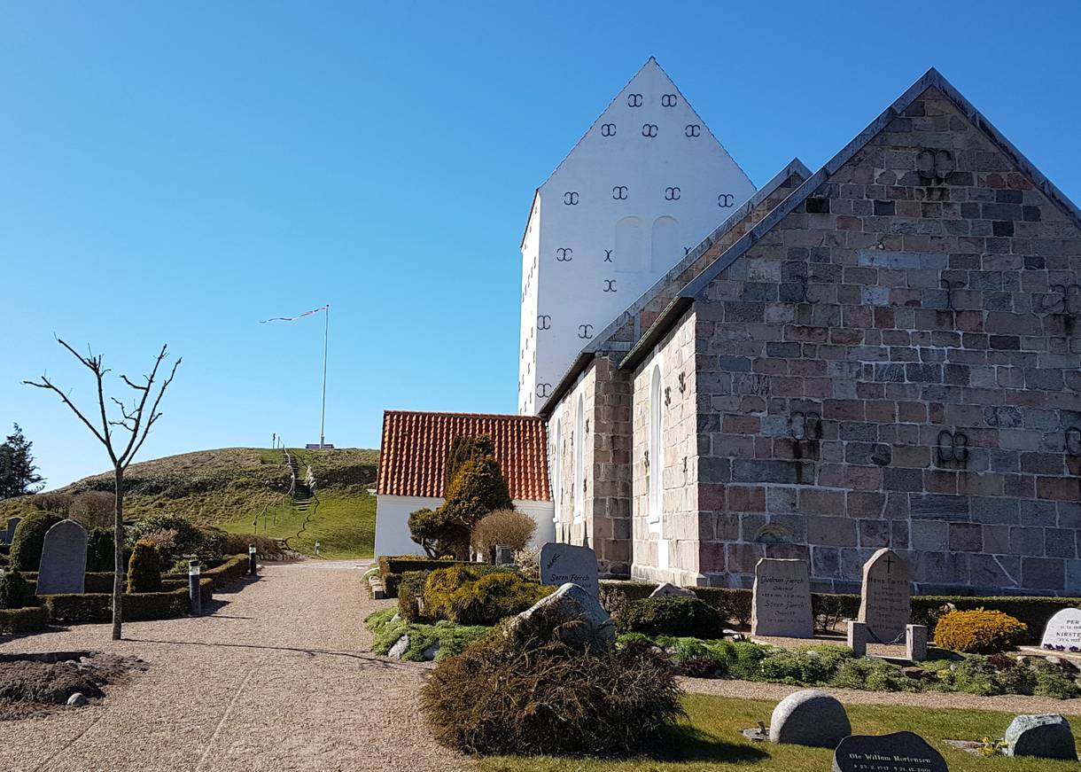 Eine graue Steinkirche mit weißem Turm, dahinter ein Grabhügel mit Fahnenmast