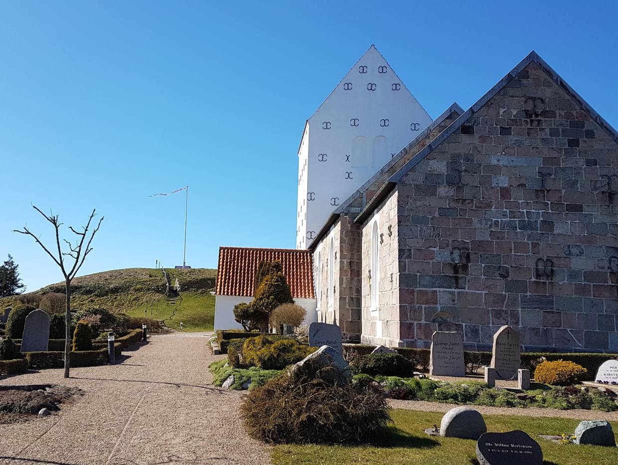 Eine graue Steinkirche mit weißem Turm, dahinter ein Grabhügel mit Fahnenmast