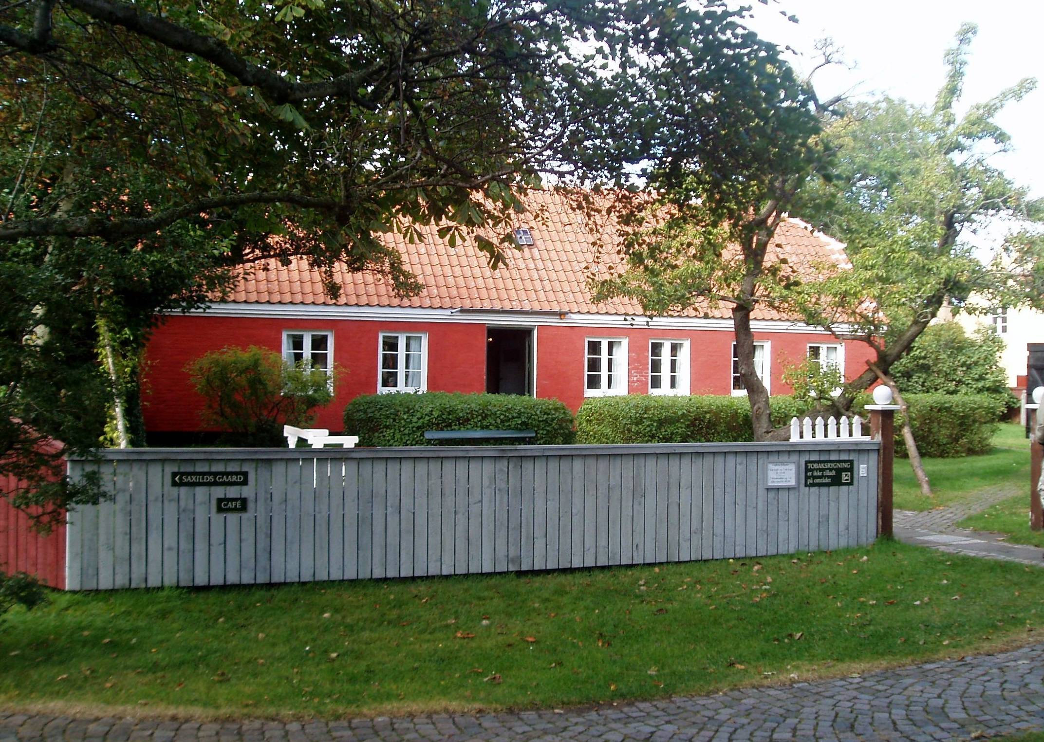 Rotes Wohnhaus in einem Garten mit Bäumen, umgeben von einem weißen Zaun