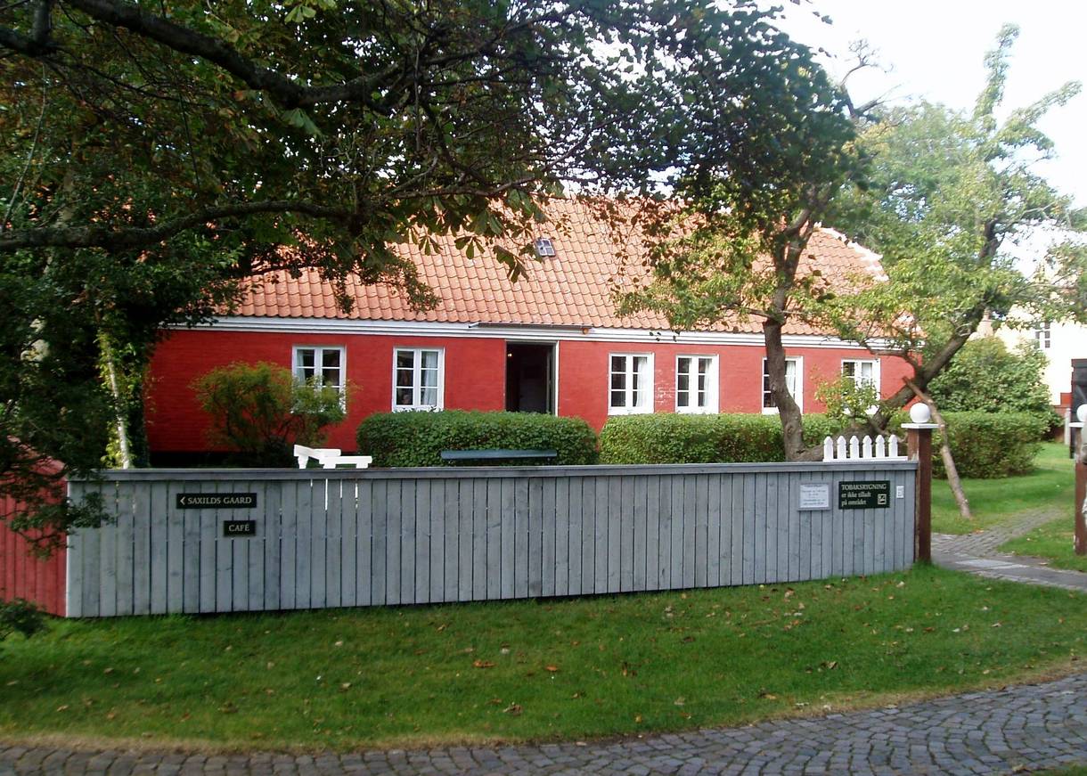 Rotes Wohnhaus in einem Garten mit Bäumen, umgeben von einem weißen Zaun