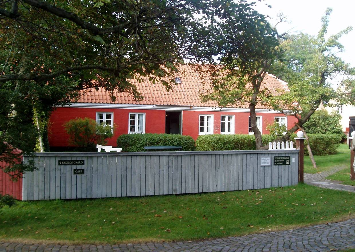 Rotes Wohnhaus in einem Garten mit Bäumen, umgeben von einem weißen Zaun