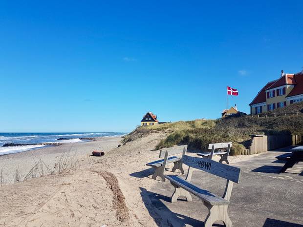 Zwei alte Sommerhäuser direkt auf einer Steilküste am Meer, davor Holzbänke und eine dänische Fahne