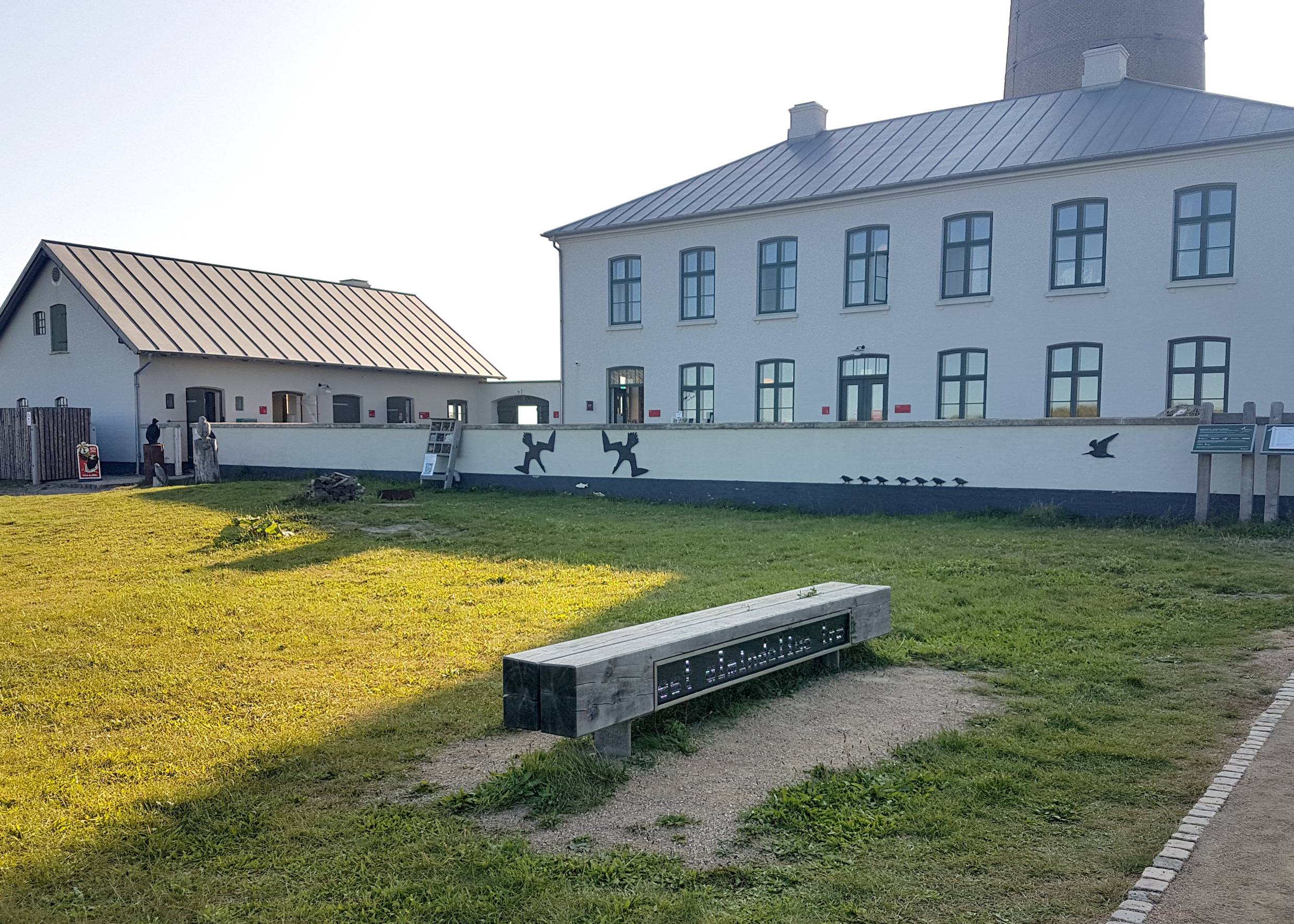 Weißes Nebengebäude des Museums, davor eine Rasenfläche mit verschiedenen Holztafeln und einer Bank