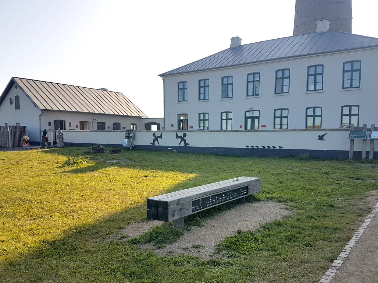 Weißes Nebengebäude des Museums, davor eine Rasenfläche mit verschiedenen Holztafeln und einer Bank