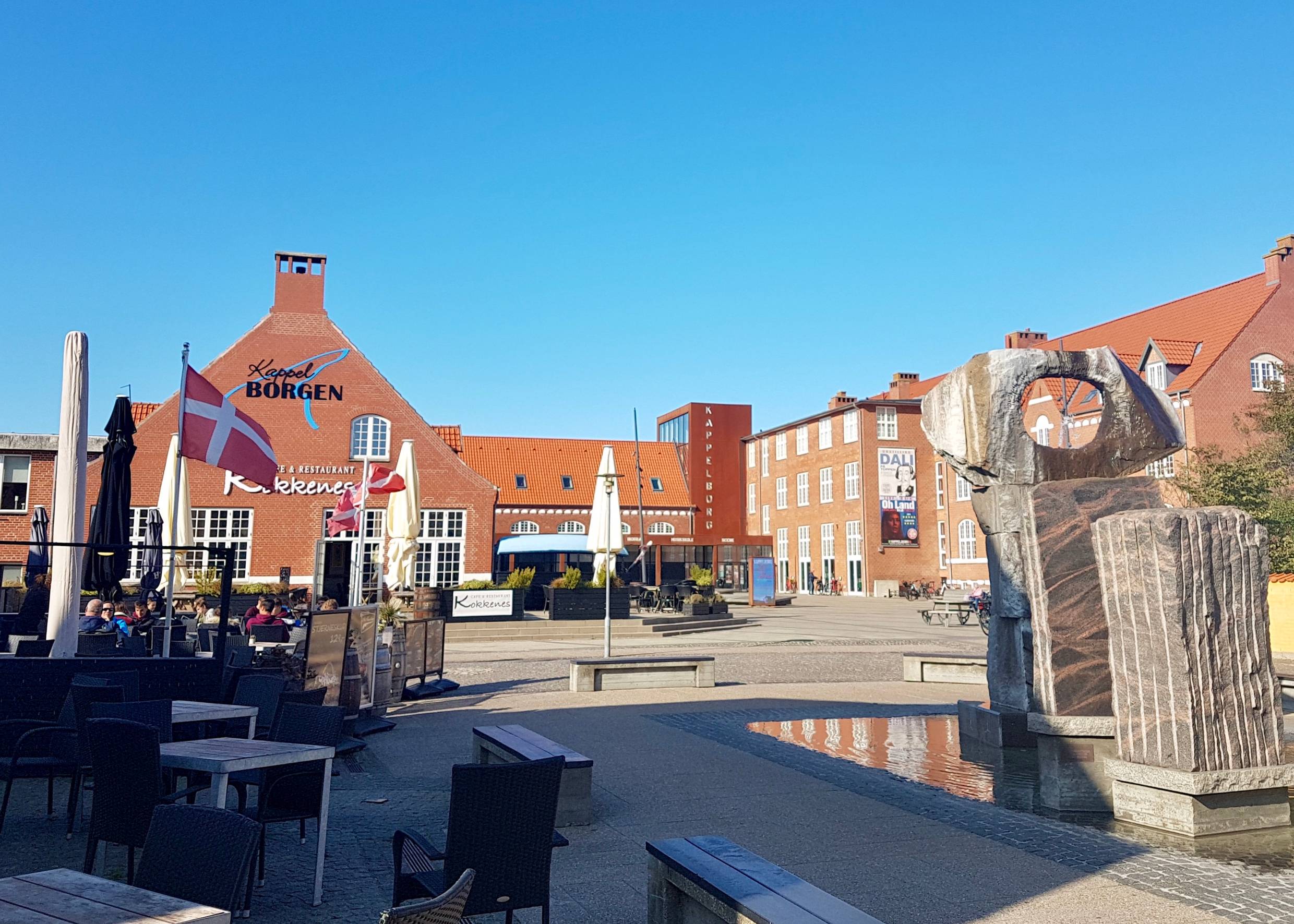 Platz an der Füßgängerzone mit einer Skulptur, Restaurantterrassen und einem Veranstaltungssaal im Hintergrund