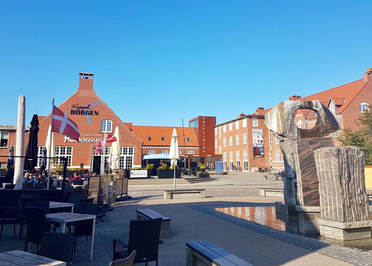 Platz an der Füßgängerzone mit einer Skulptur, Restaurantterrassen und einem Veranstaltungssaal im Hintergrund