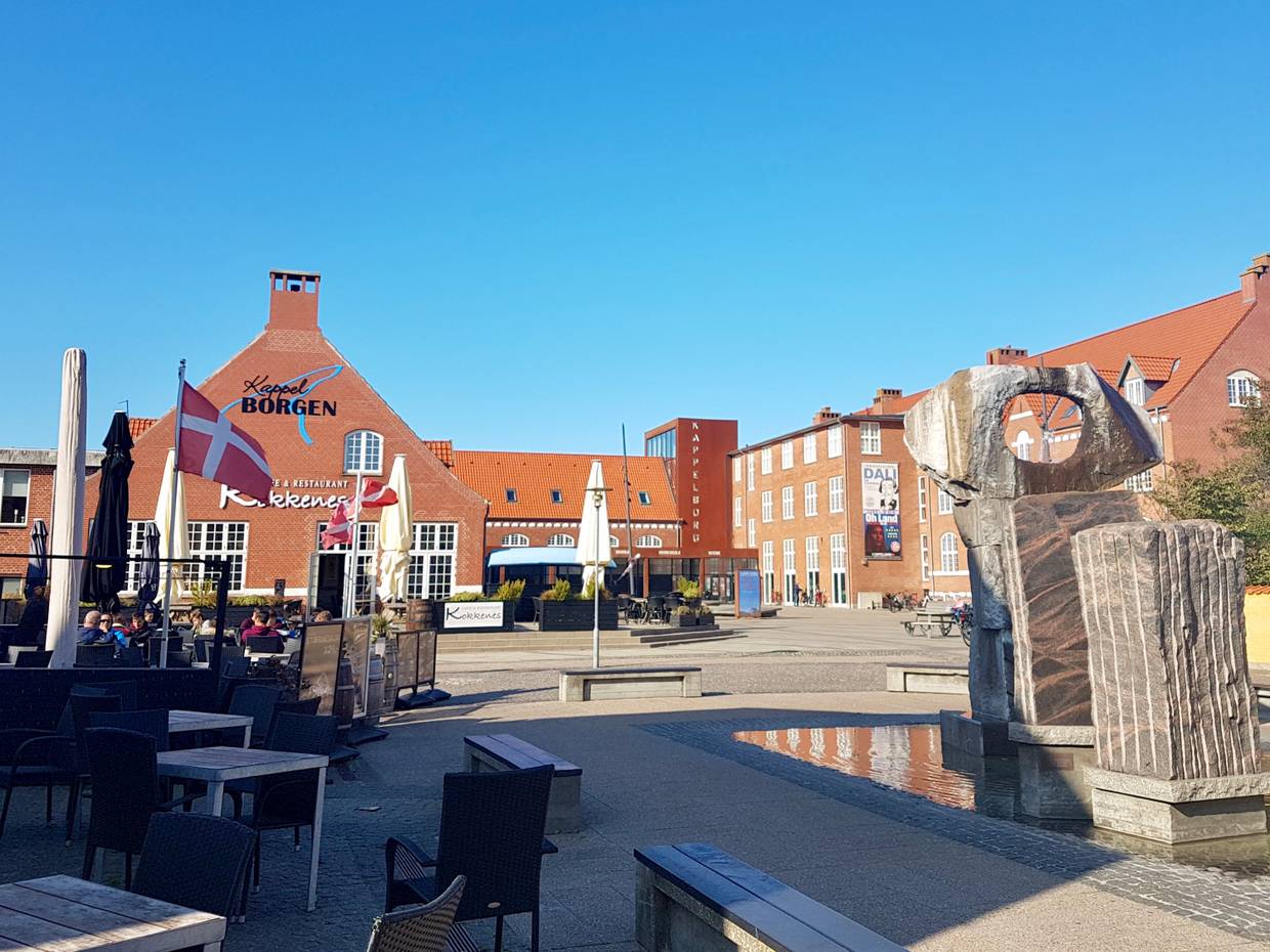 Platz an der Füßgängerzone mit einer Skulptur, Restaurantterrassen und einem Veranstaltungssaal im Hintergrund