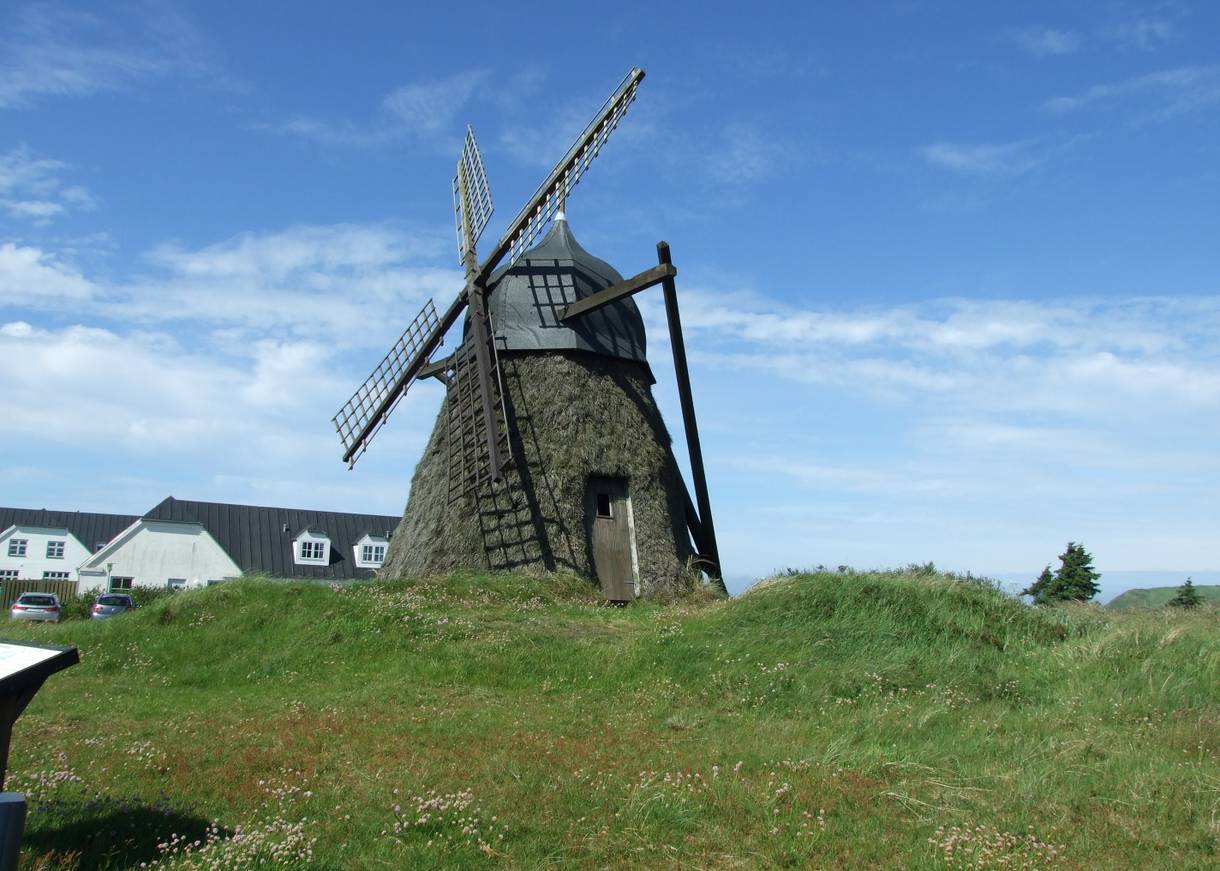 kleine Windmühle, die mit Reisig verkleidet ist, dahinter ein weißer Hof