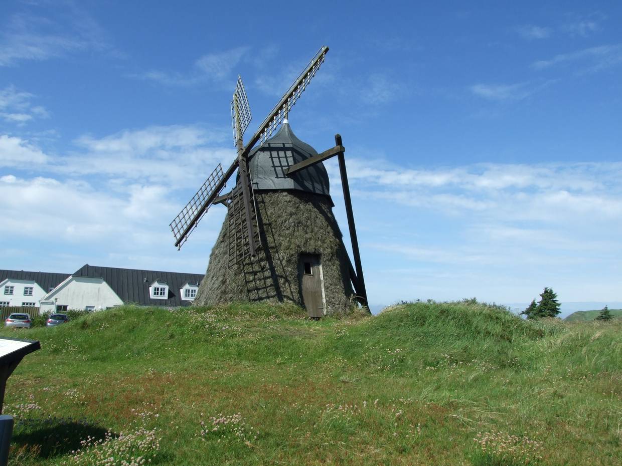 kleine Windmühle, die mit Reisig verkleidet ist, dahinter ein weißer Hof