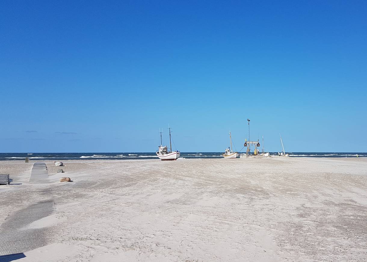 Blick vom Parkplatz auf einen breiten Strand mit zwei Booten auf dem Sand
