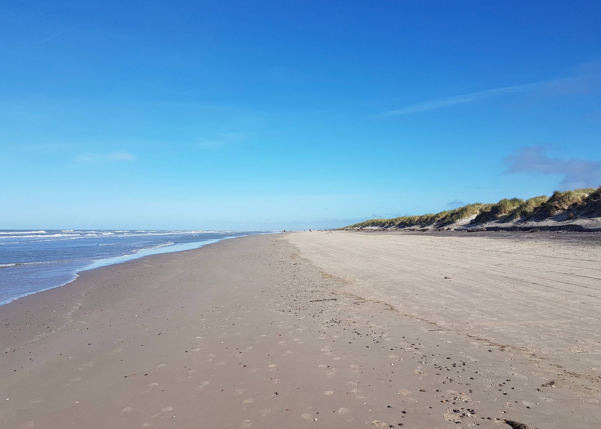 Fast menschenleerer, breiter  Strand bei Sonne, umgeben von Dünen