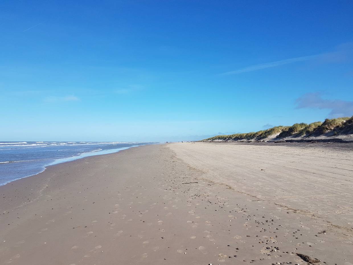 Fast menschenleerer, breiter  Strand bei Sonne, umgeben von Dünen