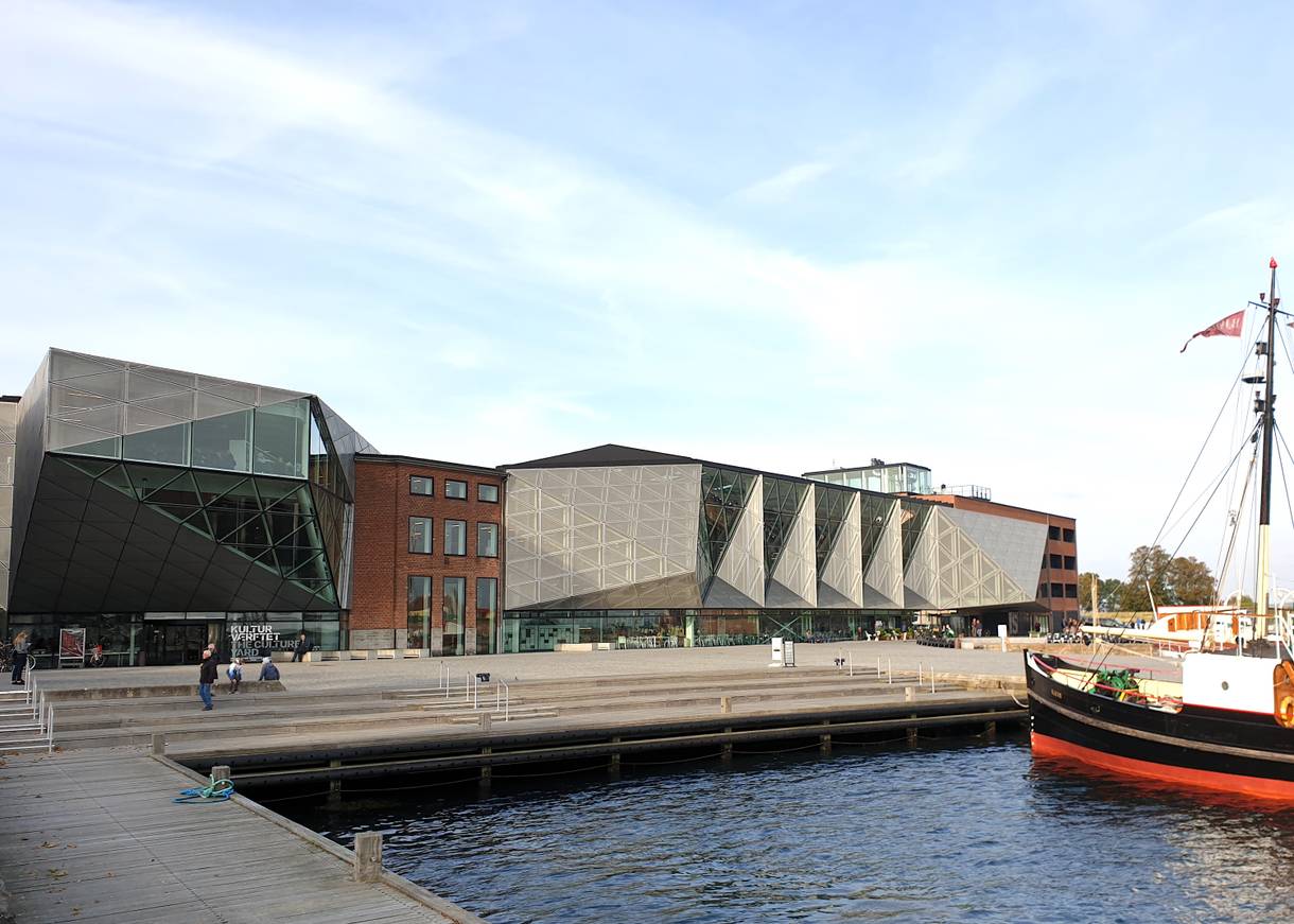 Großes, modernes Kulturgebäude an einer Uferpromanade, im Wasser ein kleines Dampfschiff