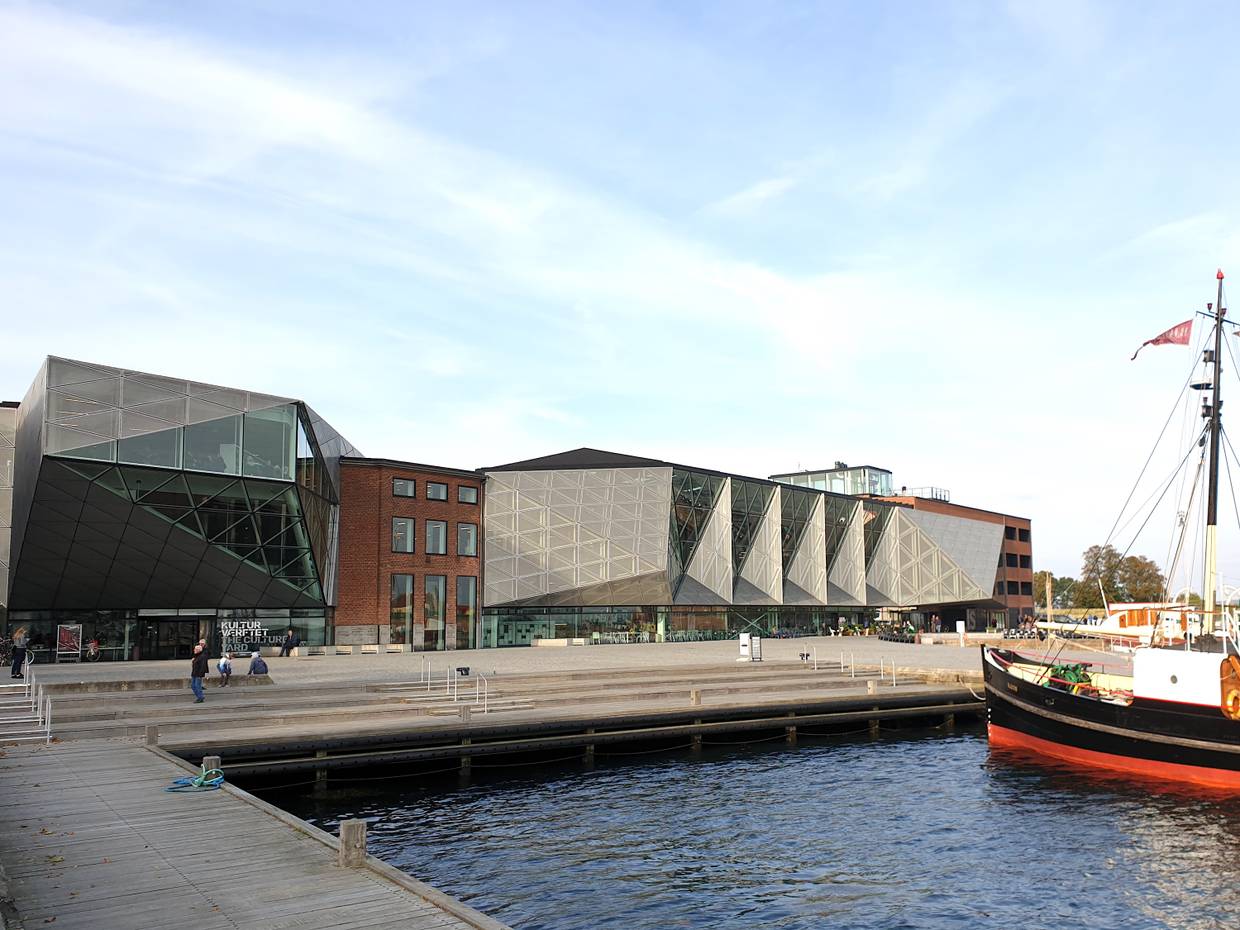 Großes, modernes Kulturgebäude an einer Uferpromanade, im Wasser ein kleines Dampfschiff