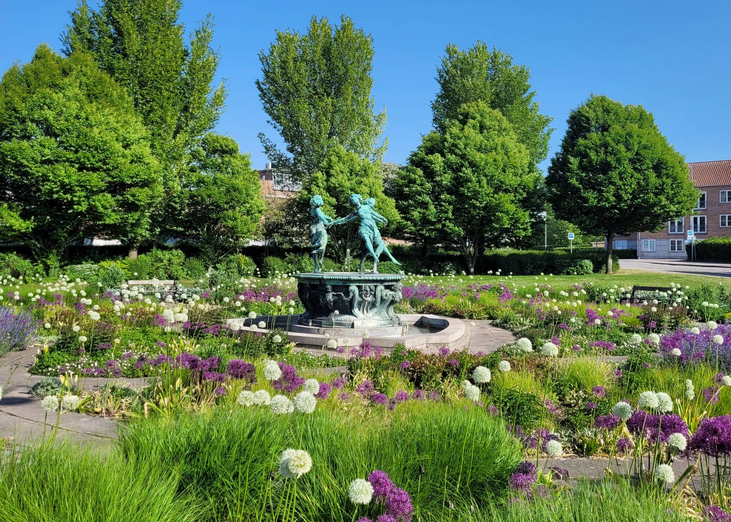 Ein Platz mit vielen Blumen, in der Mitte ein Brunnen mit einer Gruppe tanzender Frauen