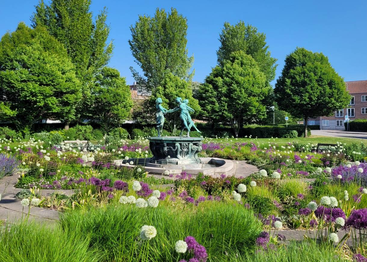 Ein Platz mit vielen Blumen, in der Mitte ein Brunnen mit einer Gruppe tanzender Frauen