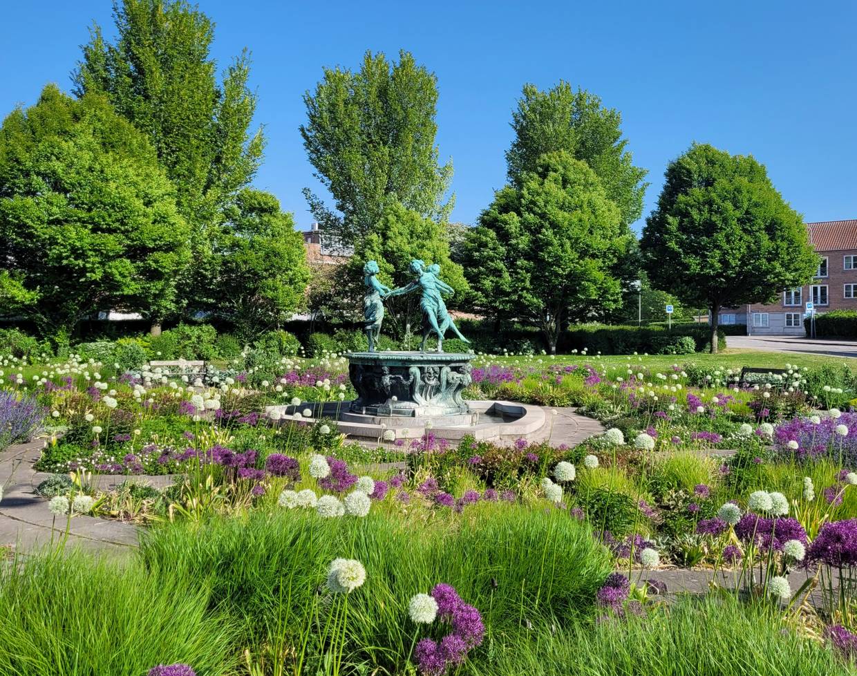 Ein Platz mit vielen Blumen, in der Mitte ein Brunnen mit einer Gruppe tanzender Frauen