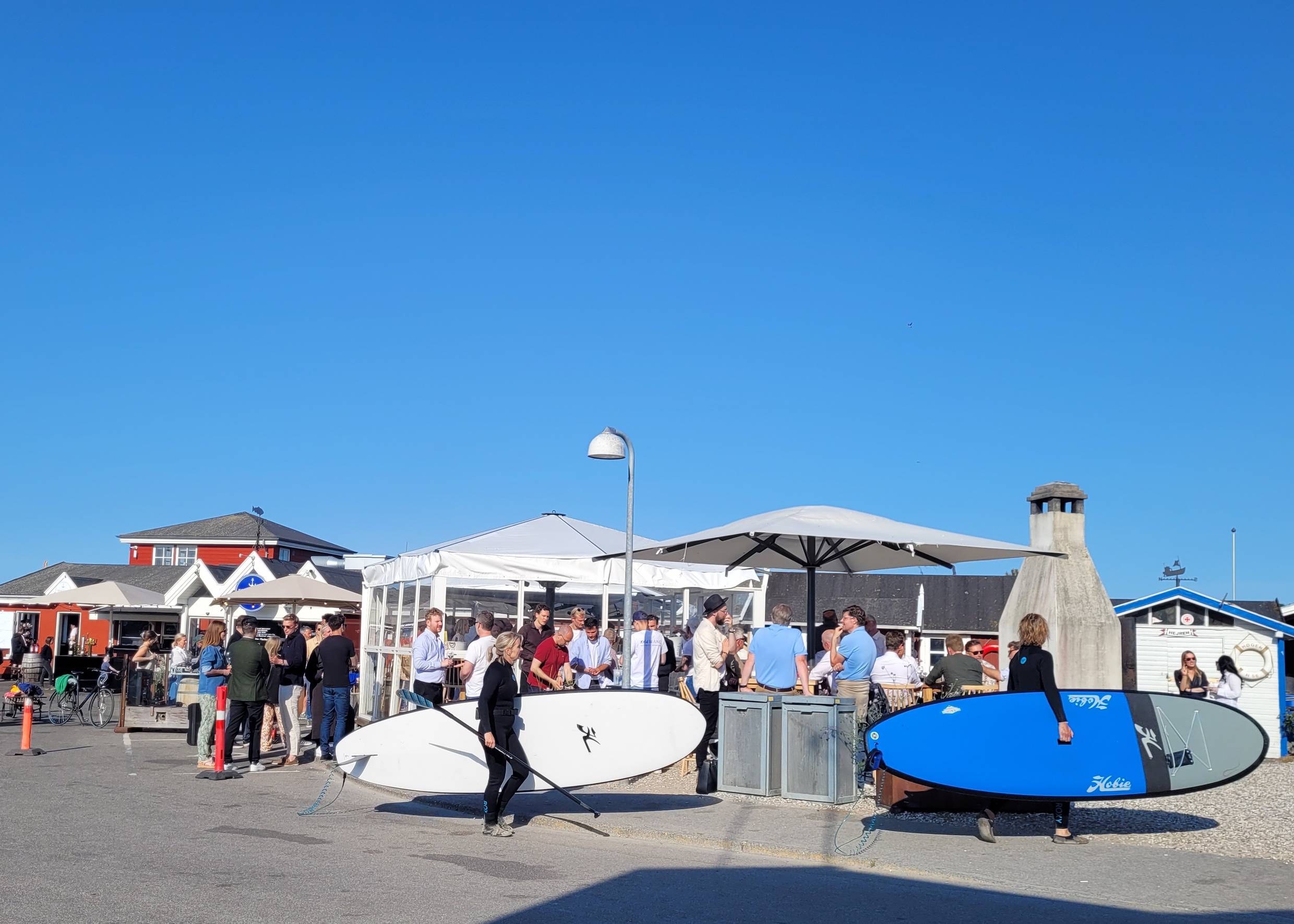 Hafenlokal mit vielen Gästen, davor zwei Frauen mit einem Surfboard unter dem Arm