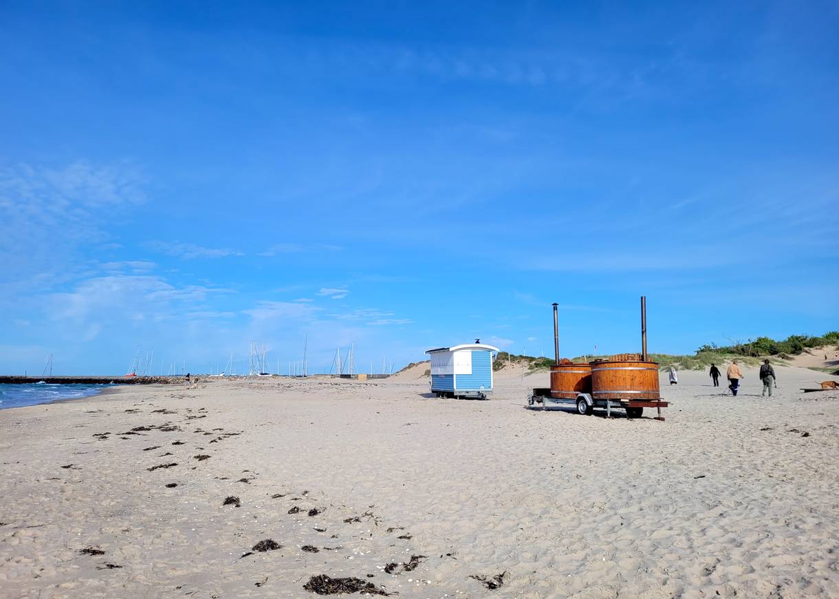 Ein Anhänger mit zwei Holztubs und ein blauber Bauwagen auf einem breiten Sandstrand
