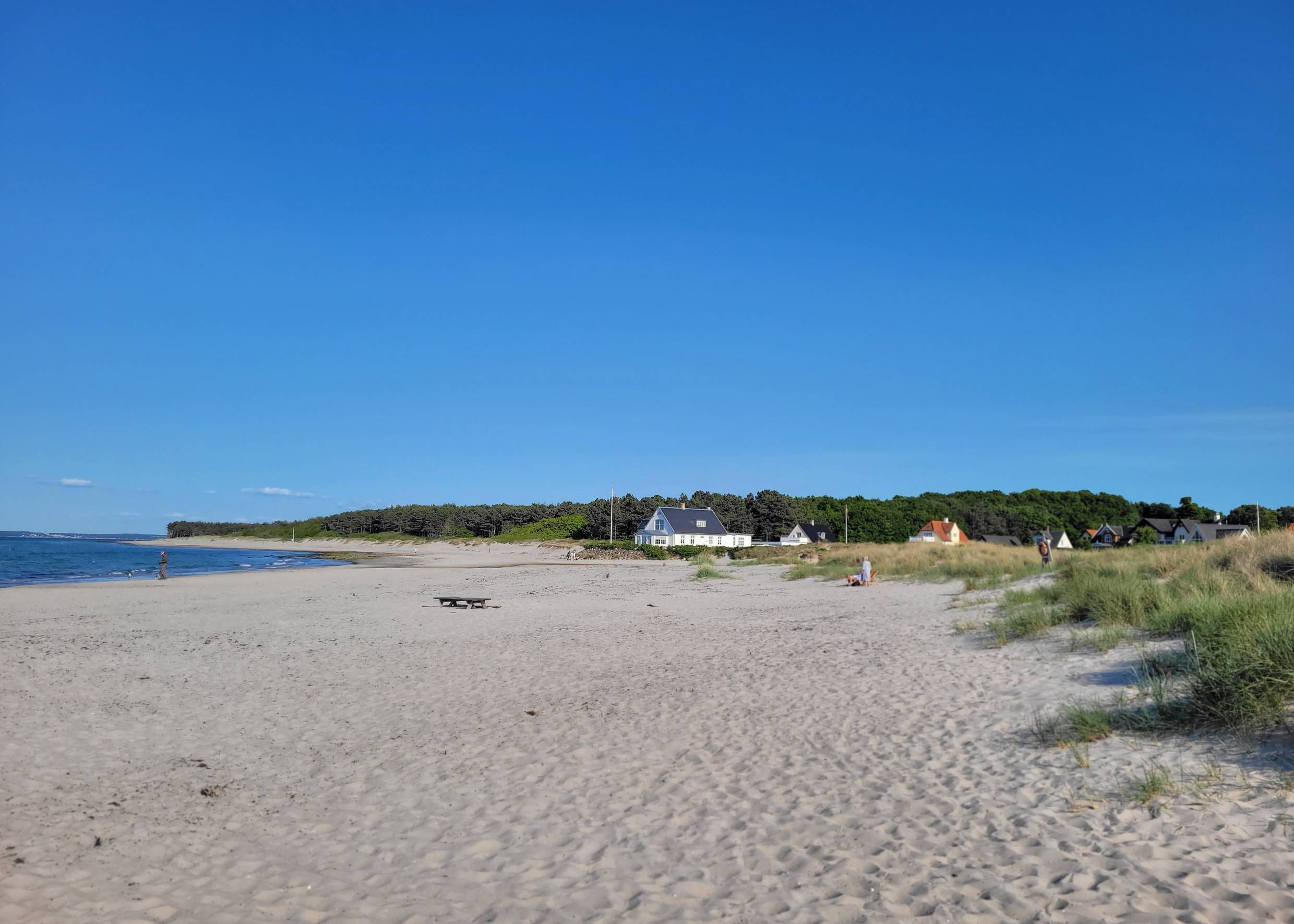 Breiter Sandstrand, im Hintergrund Wald und ein paar Häuser mit Meerblick