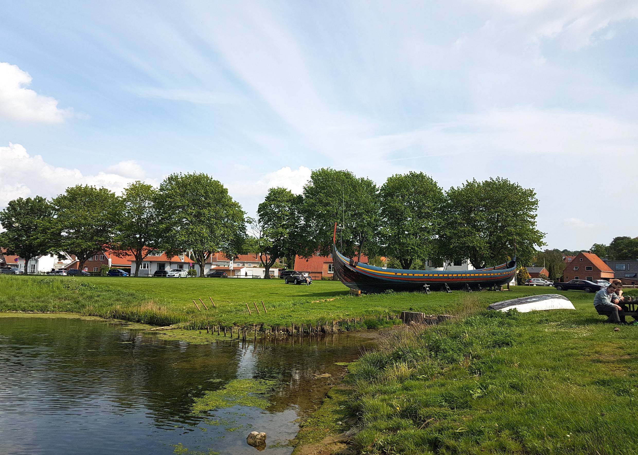 Farbiges Holzboot auf einer Wiese, dahinter Wohnhäuser