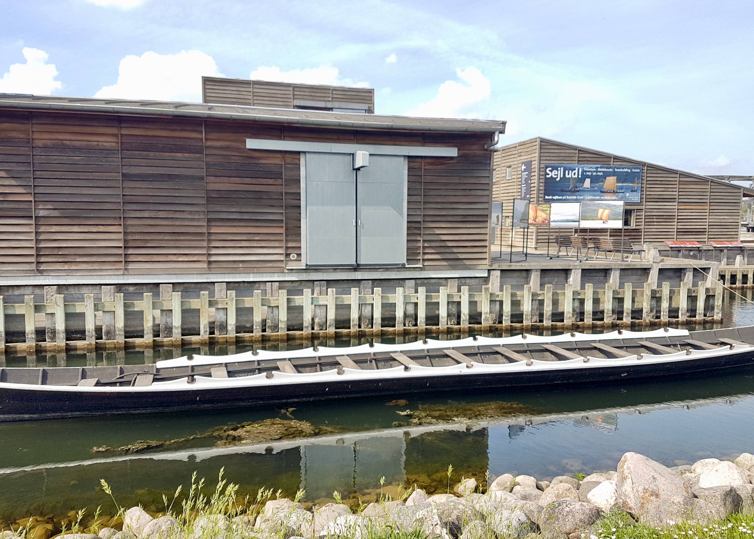 Flacher Holzbau am Wasser, davor ein langes, schmales Schiff 