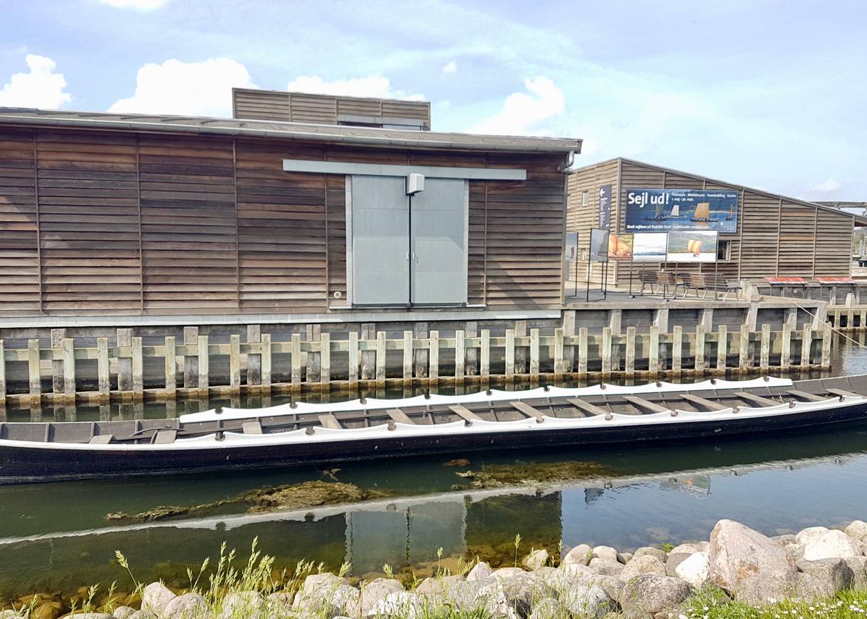 Flacher Holzbau am Wasser, davor ein langes, schmales Schiff 
