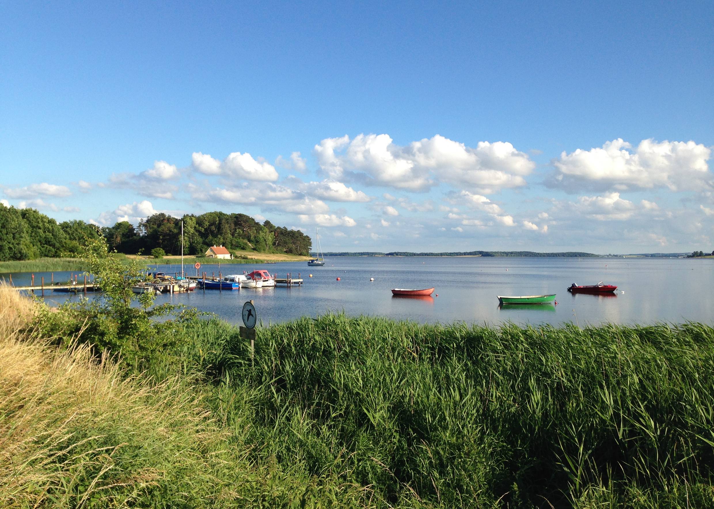 Bootssteg in einer grün umrandeten Bucht, im Wasser bunte Boote unter blauem Himmel