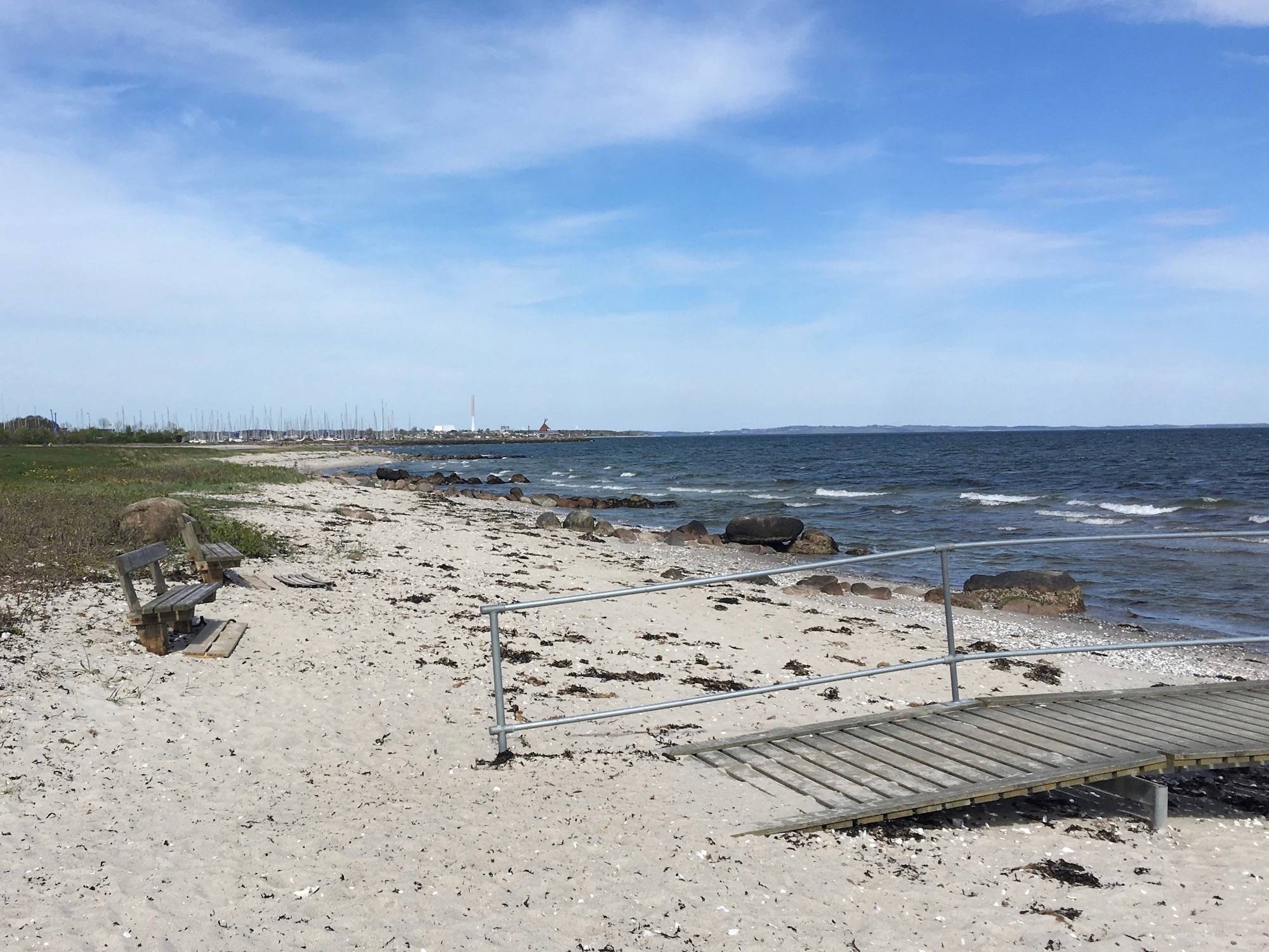 Sandstrand mit trockenem Seetang, ein Steg auf dem Sand führt zum Wasser