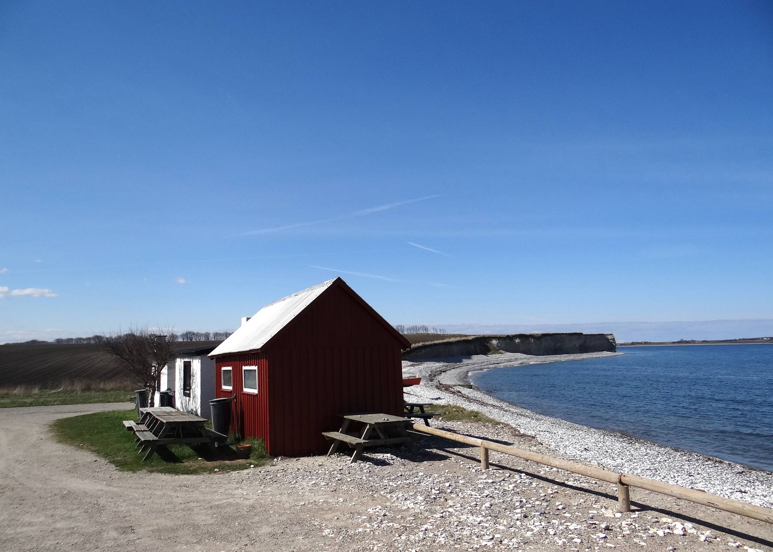 Rote Fischerhütte mit Picknickbänken vor einer sehr steinigen Küste am Meer