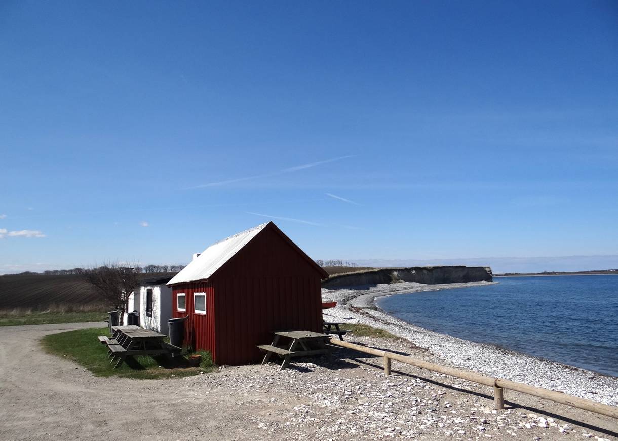 Rote Fischerhütte mit Picknickbänken vor einer sehr steinigen Küste am Meer
