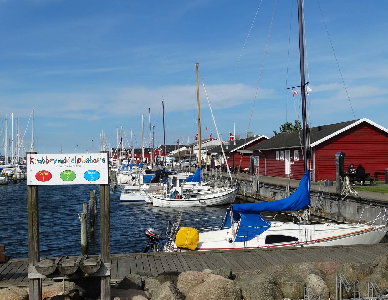 Segelboote an der Hafenmauer, dahiner rote Fischerhütten, im Vordergrund ein Holzsteg