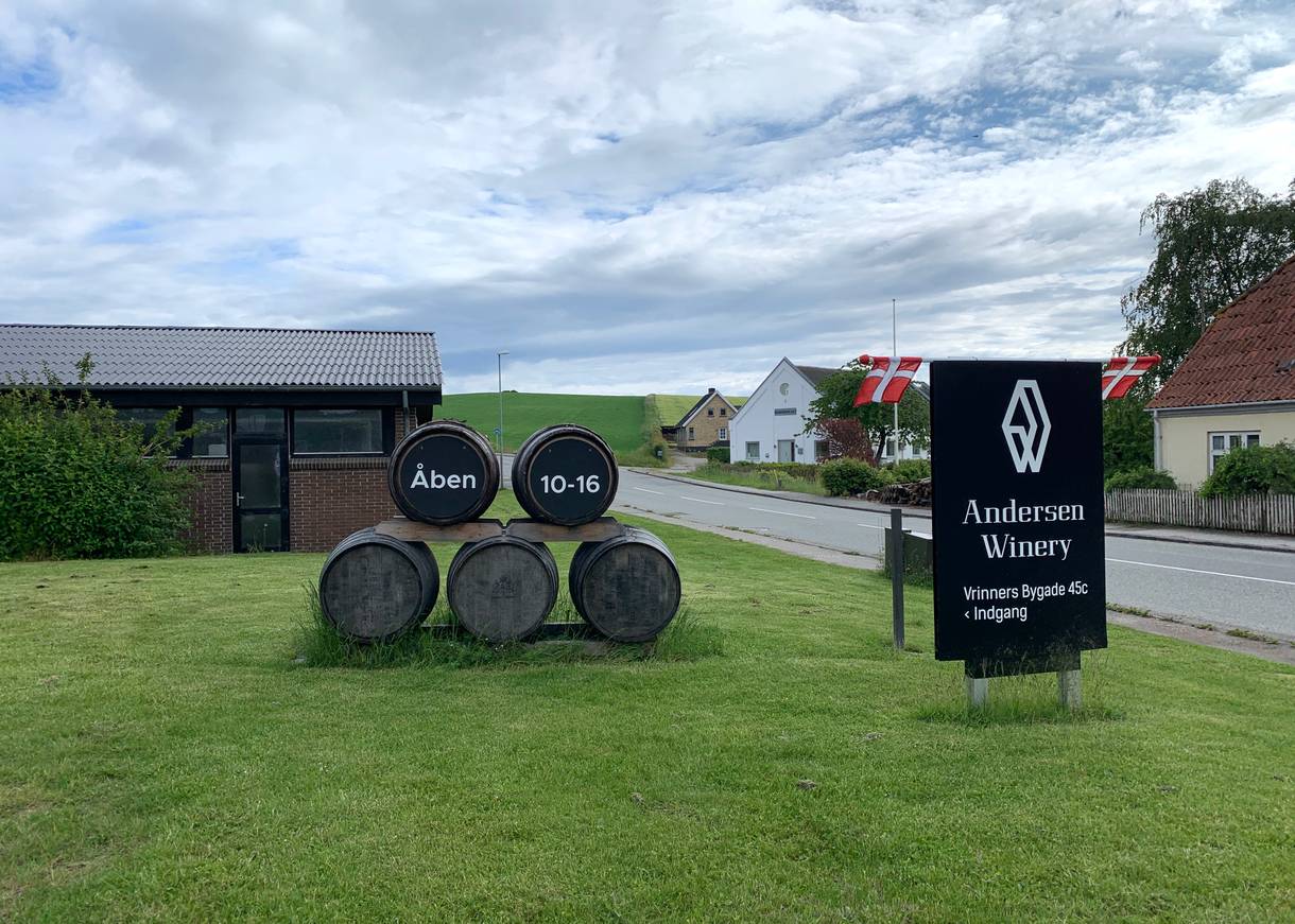 Gestapelte Weinfässer auf der Wiese vor einem Haus mit Öffnungszeiten zum Hofladen der Winery
