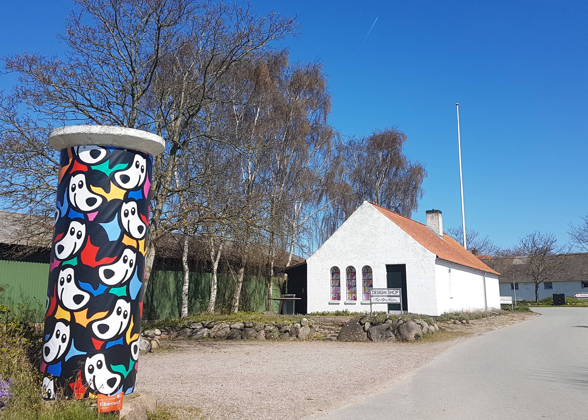 Litfaßsäule mit bunten Figuren an einer Dorfstraße