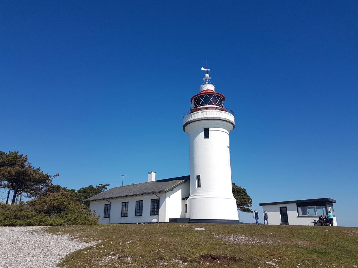 Kleiner weißer Leuchtturm mit Nebengebäude, davor Kies und Rasen, blauer Himmel über Sletterhage Fyr