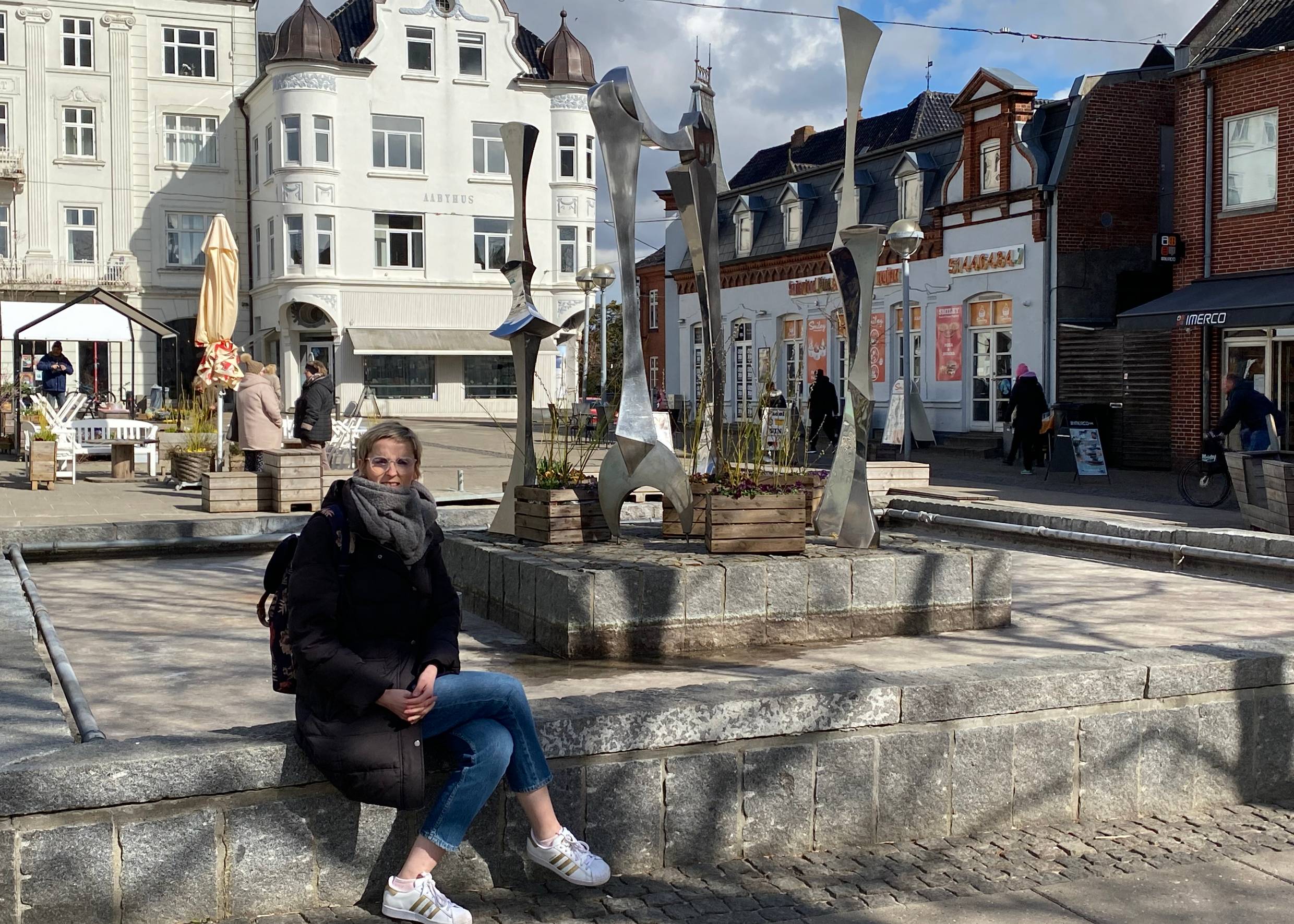 Brunnen mit Kunstinstallation vor einem weißen Bürgerhaus, eine Frau sitzt am Brunnen