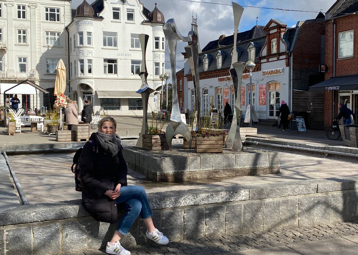 Brunnen mit Kunstinstallation vor einem weißen Bürgerhaus, eine Frau sitzt am Brunnen