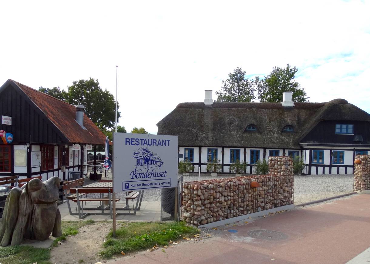 Fachwerkhaus mit Parkplatz davor, links ein Kiosk und eine Skuptur 
