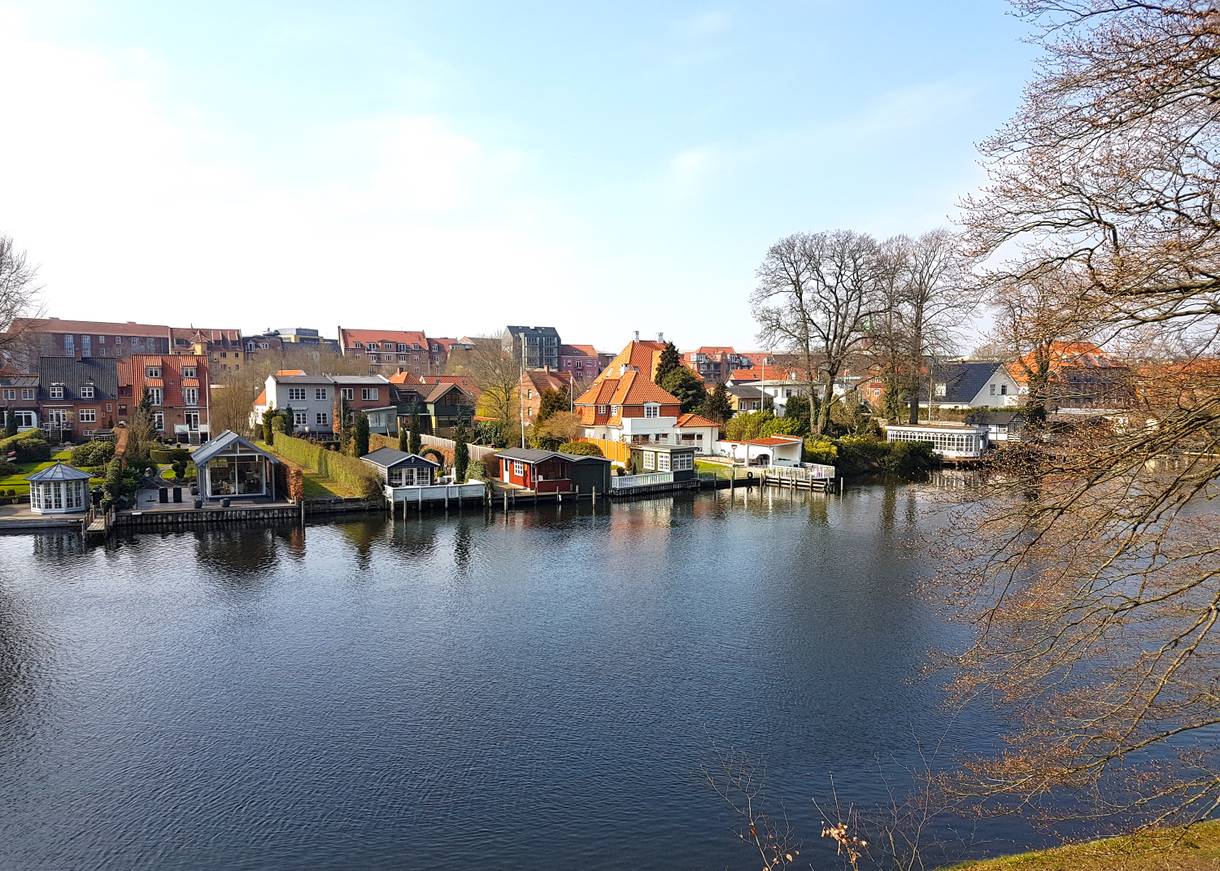 Blick über einen Fluss, am gegenüberliegenden Ufer stehen Häuser direkt am Wasser