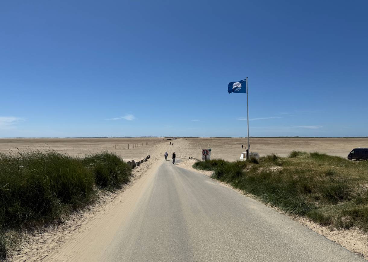 Asphaltierter Weg mit einer gehissten blauen Flagge und einer breiten Strand