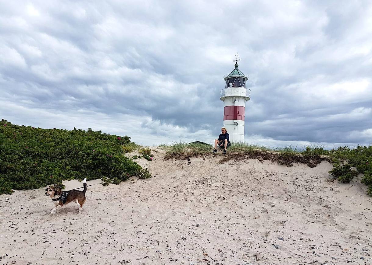 Kleiner rot-weißer Leuchtturm, davor sitzt eine Frau im Sand mit einem Hund