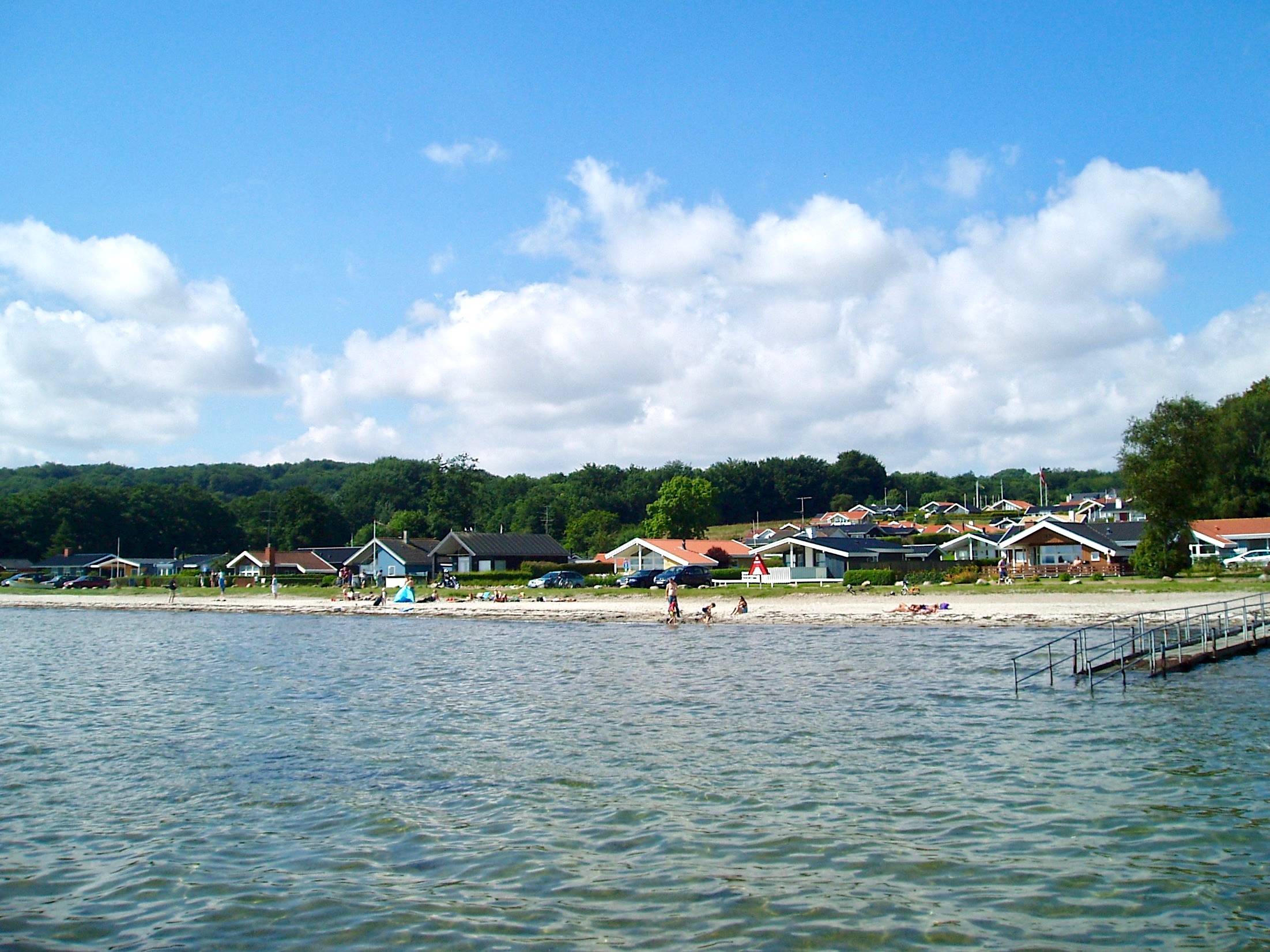Blick vom Wasser auf einen schmalen Naturstrand mit Badegästen, dahinter ferienhäuser