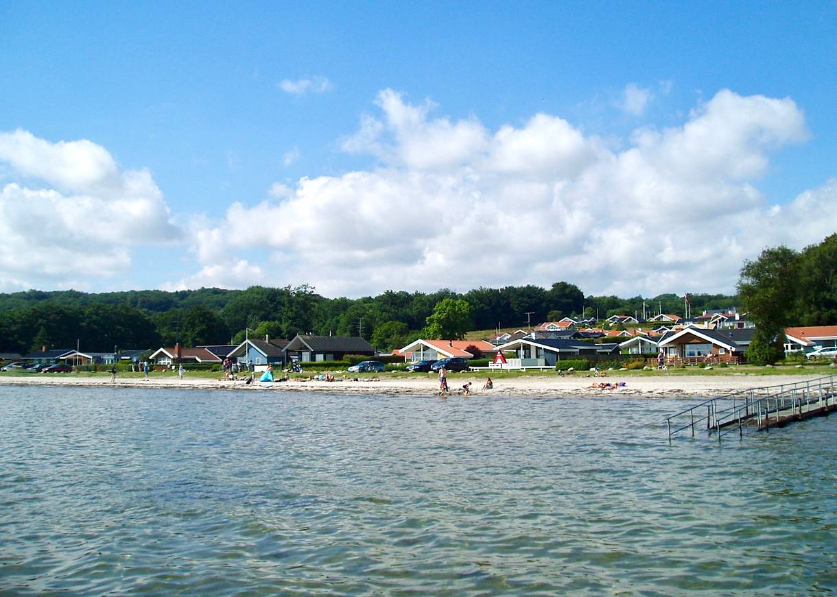 Blick vom Wasser auf einen schmalen Naturstrand mit Badegästen, dahinter ferienhäuser