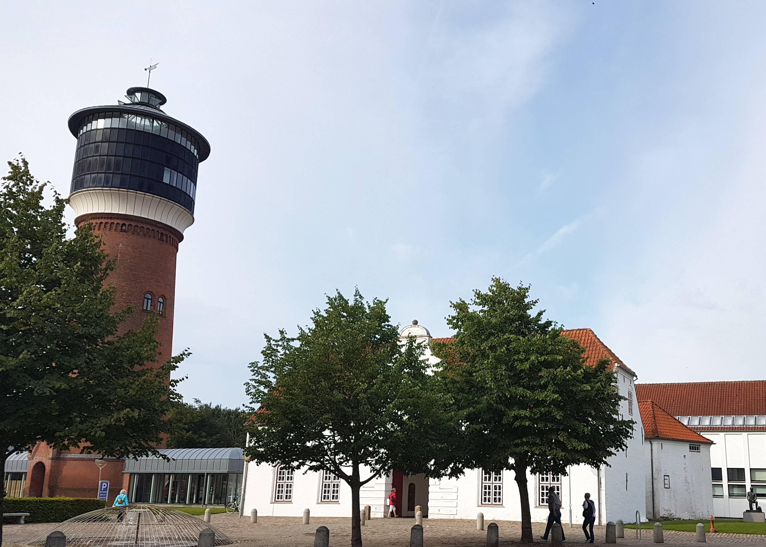 Weißes Gebäude mit einem offenen Durchgang in der Mitte, links daneben ein Wasserturm