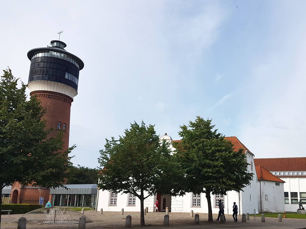 Weißes Gebäude mit einem offenen Durchgang in der Mitte, links daneben ein Wasserturm
