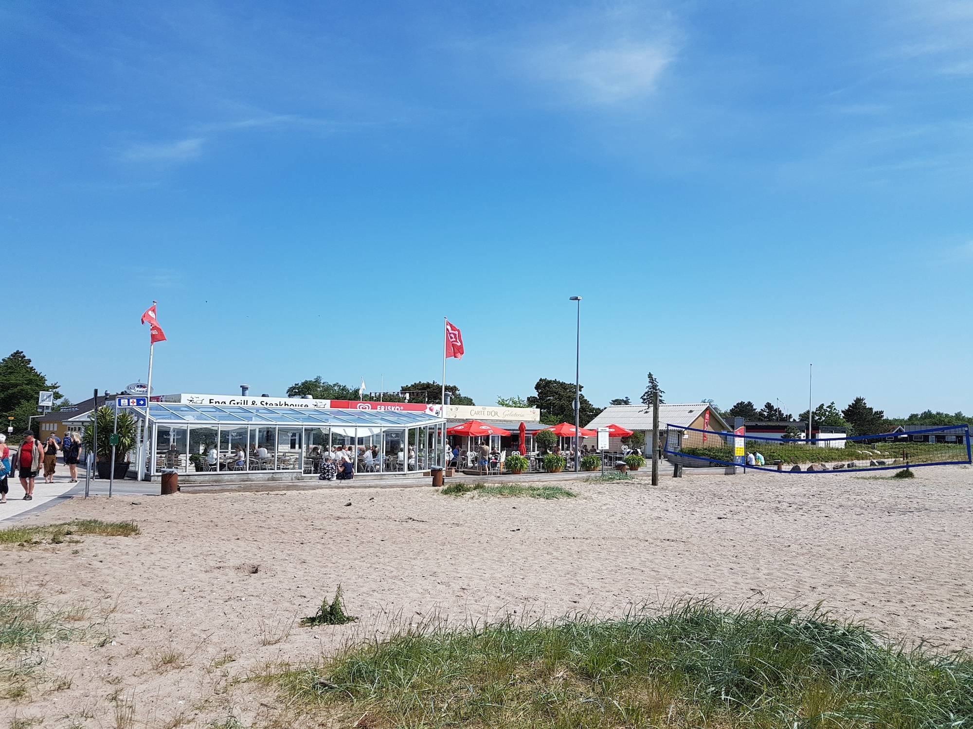 Restaurant mit vielen Gästen am Strand, davor Sand und ein Beachvolleyballfeld