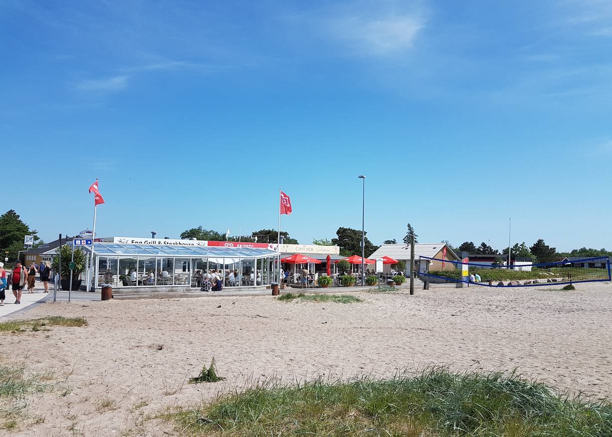 Restaurant mit vielen Gästen am Strand, davor Sand und ein Beachvolleyballfeld