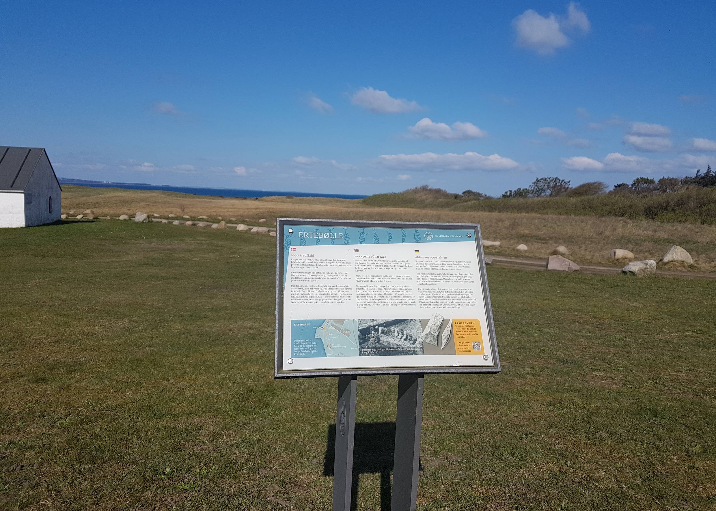 Info-Schild vor einer Wiese, am Bildrand ein kleines, weißes Haus